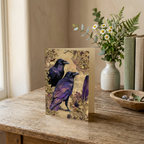 Ravens & Gilded Pages portrait greeting card on a wooden table beside a vase of flowers.